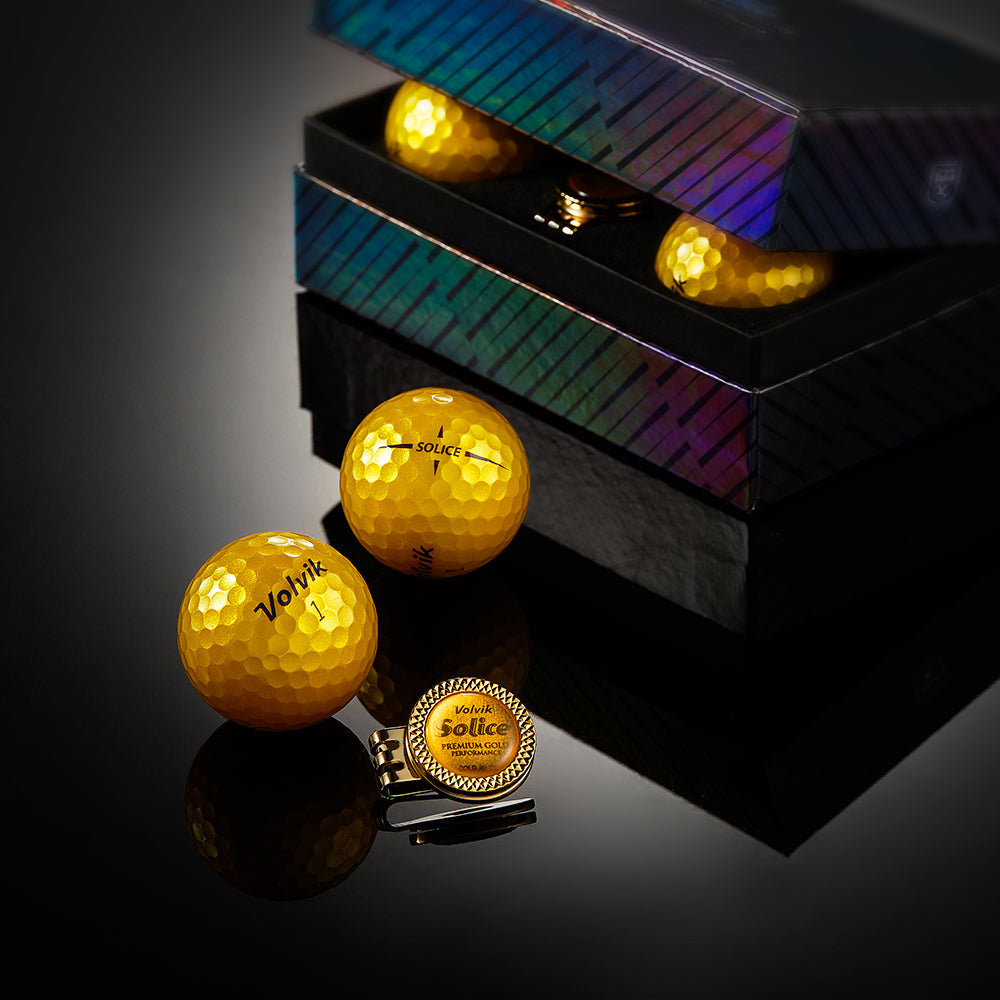 Two gold Volvik golf balls with a ball marker on a reflective black surface