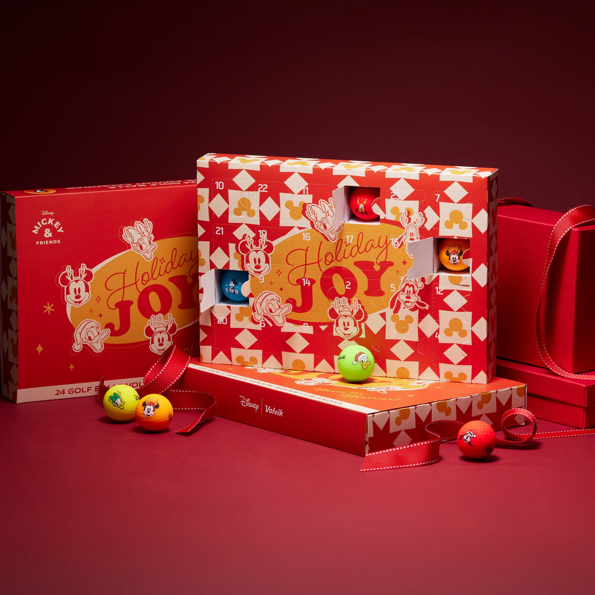 A Disney-themed holiday advent box set displayed on a dark red studio background. The set includes a large central advent calendar box with red and white patterned sides, a smaller matching box behind it, a red gift bag, and a few small colorful advent balls placed in front.