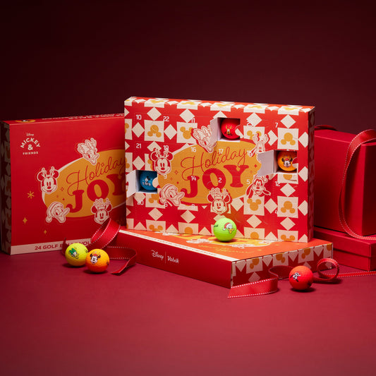 A Disney-themed holiday advent box set displayed on a dark red studio background. The set includes a large central advent calendar box with red and white patterned sides, a smaller matching box behind it, a red gift bag, and a few small colorful advent balls placed in front.