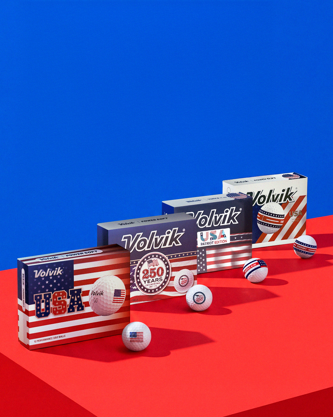 Five boxes of Volvik USA golf balls arranged diagonally on a red platform against a solid blue background. The boxes feature patriotic American flag designs in red, white, and blue, and several golf balls are scattered around them.
