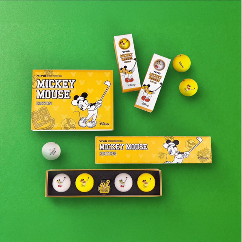 A set of golf balls with Mickey Mouse design on them, along with packaging displaying the same design, placed on a green background.